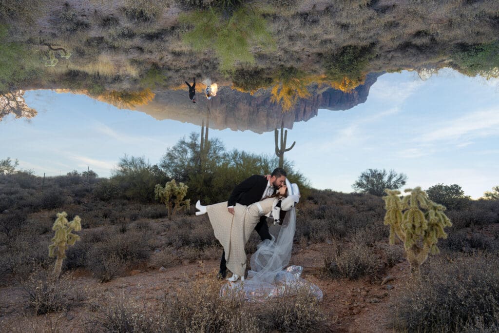 Couple eloping in Arizona kissing as he dips her in a cowgirl hat with the Superstition Mountains behind them and an artistic upside-down mountain reflection in the sky